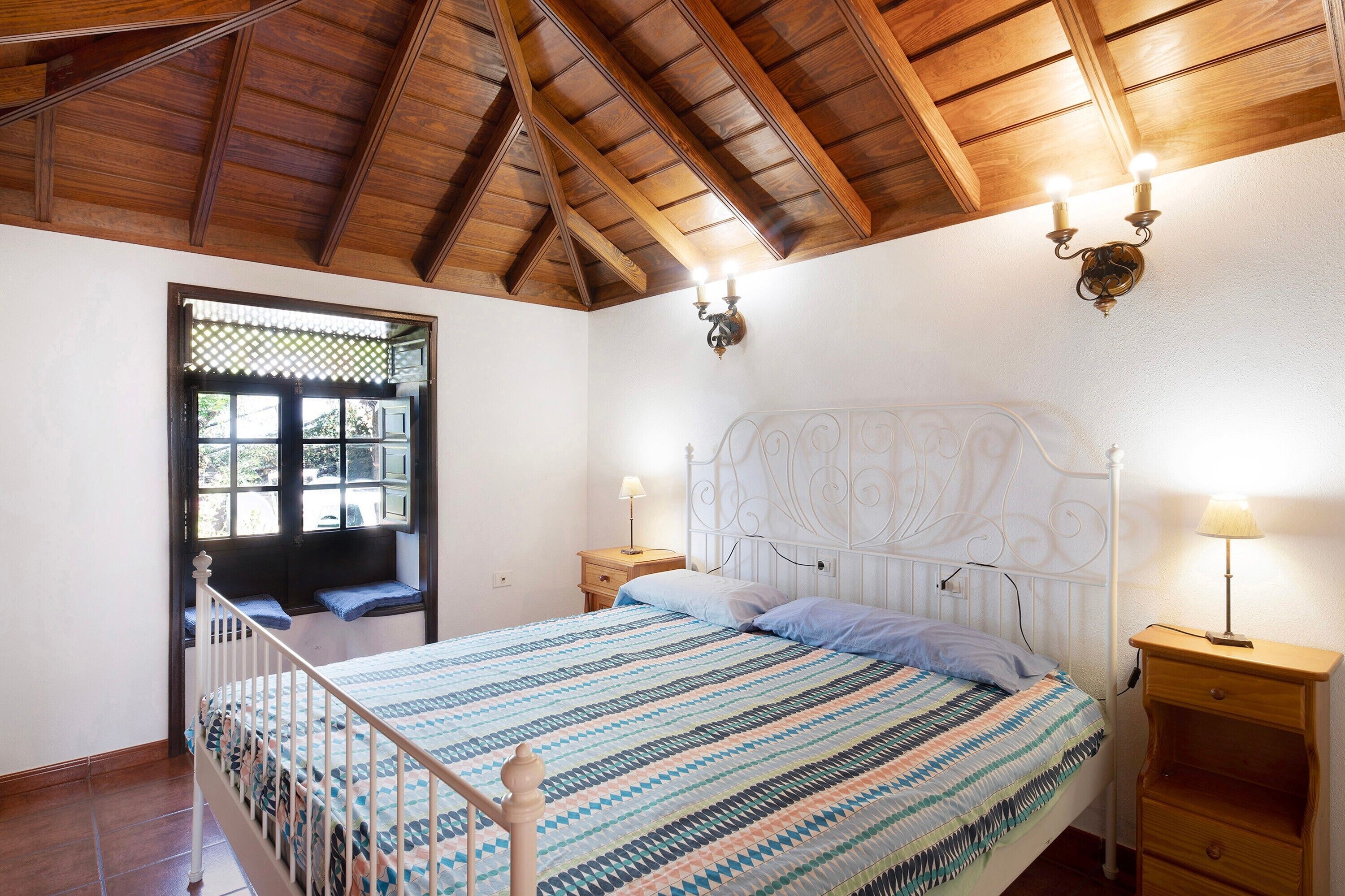 A bedroom with a wooden ceiling, a bed with a patterned bedspread, two bedside lamps, and a window with a view.