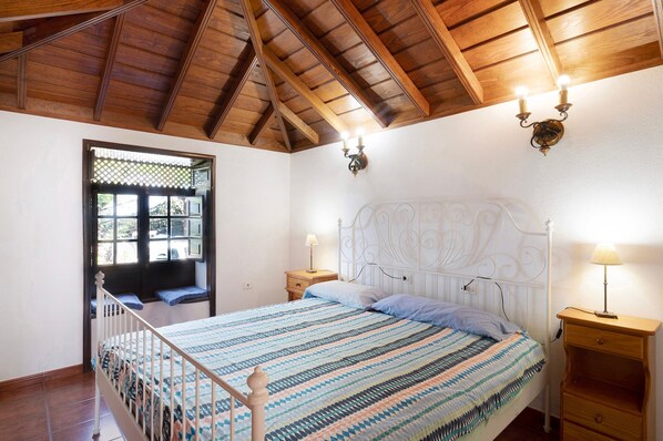 Une chambre avec un plafond en bois, un lit recouvert d’une couvre-lit à motifs, deux lampes de chevet et une fenêtre donnant sur un beau paysage.