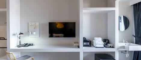 A modern hotel room with a white desk, a flat-screen TV, a small seating area, a compact refrigerator, and a bathroom with a mirror and sink.