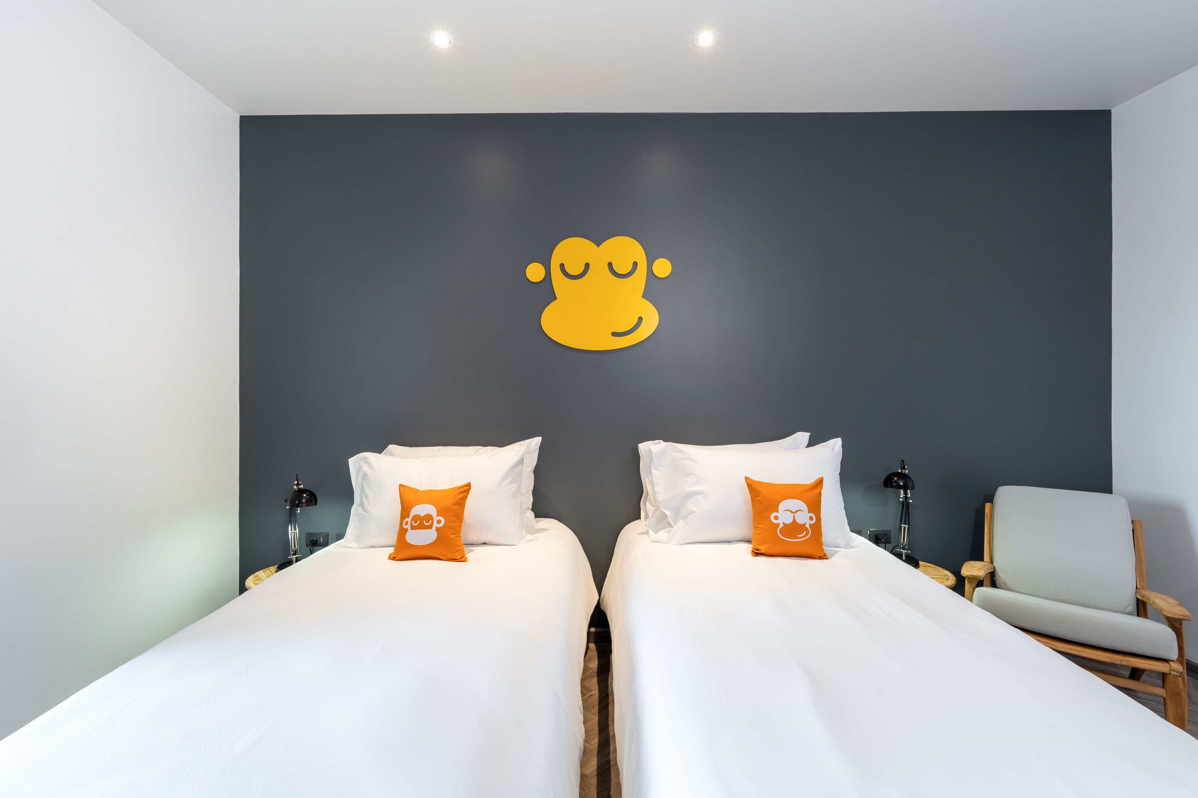 Hotelkamer met twee bedden, elk met witte lakens en oranje kussens, een grijze accentmuur met een geel logo, een grijze fauteuil en een nachtkastje met een lamp.