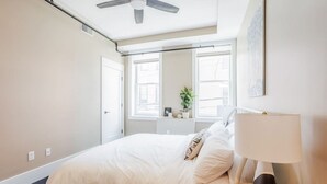 Un dormitorio con cama, lámpara de noche, ventana y ventilador de techo.