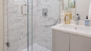 Un baño moderno con ducha envuelta en cristal, paredes de mármol y tocador con artículos de aseo.