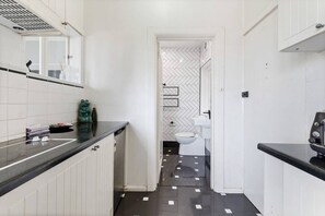 Un pasillo estrecho con una zona de cocina a un lado y un baño al otro. La cocina tiene armarios blancos y una encimera negra. El baño cuenta con un inodoro blanco y un salpicadero con estampado de espiga.