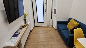 A narrow hallway with a flat-screen TV on the left, a blue sofa with a yellow pillow on the right, and a small table with yellow chairs. - PALMHOUSE Salerno center with jacuzzi, separate entrance near Metro. (Salerno)