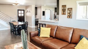 A modern living room with a brown leather sofa, a dining area with a wooden table and chairs, and a kitchen area with a white countertop and backsplash. - Amazing custom new home with modern amenities for a memorable and comfortable stay. (Moab)