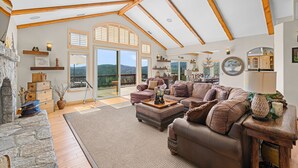 Un vaste salon doté d’un plafond haut, de poutres en bois, d’une cheminée en pierre et d’une grande fenêtre donnant sur un paysage magnifique.