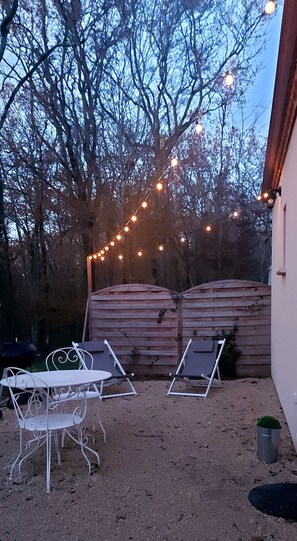 A patio with two white metal chairs and a table, surrounded by a wooden fence and trees. - Cottage "Romantique" (Saint Chamassy)