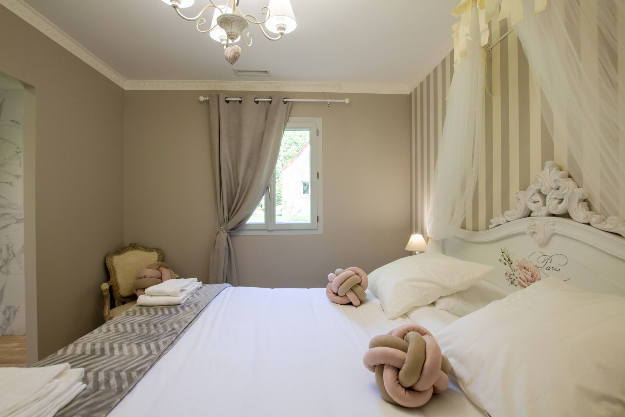 A bedroom with a bed, curtains, a chair, and a chandelier.