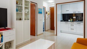 A modern living room with a TV, a white cabinet with glass doors, a kitchen area with white cabinets, and a wooden door.