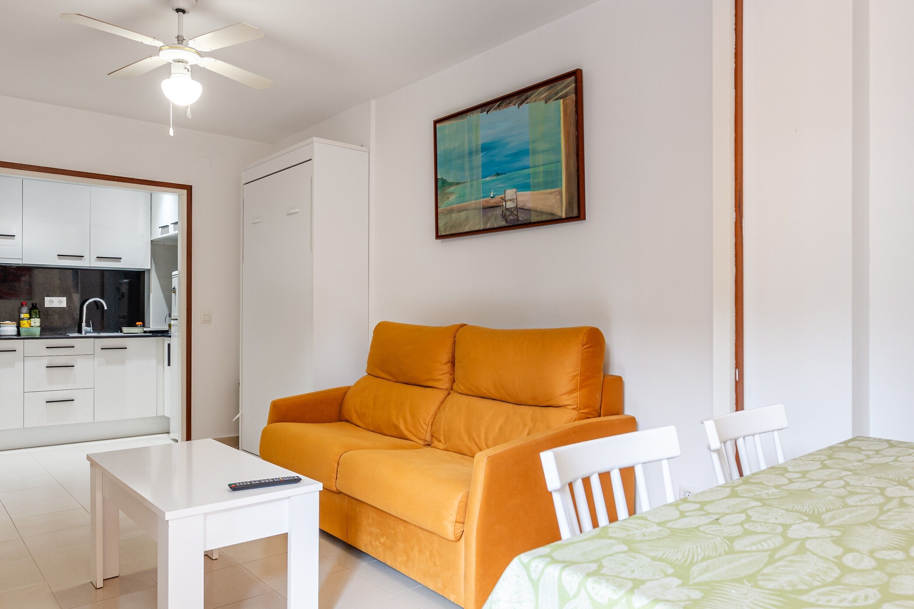 Una habitación con ventilador de techo blanco, una mesa blanca, un sofá naranja y una colcha estampada verde, además de un cuadro con escena de playa.