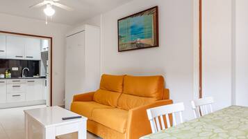 Una habitación con ventilador de techo blanco, una mesa blanca, un sofá naranja y una colcha estampada verde, además de un cuadro con escena de playa.