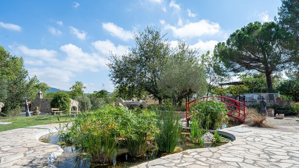 A stone pathway leading to a pond with plants, a red bridge, and trees in a park setting. - Holiday Home "Horrio" with Mountain View, Shared Pool & Wi-Fi (Querol)