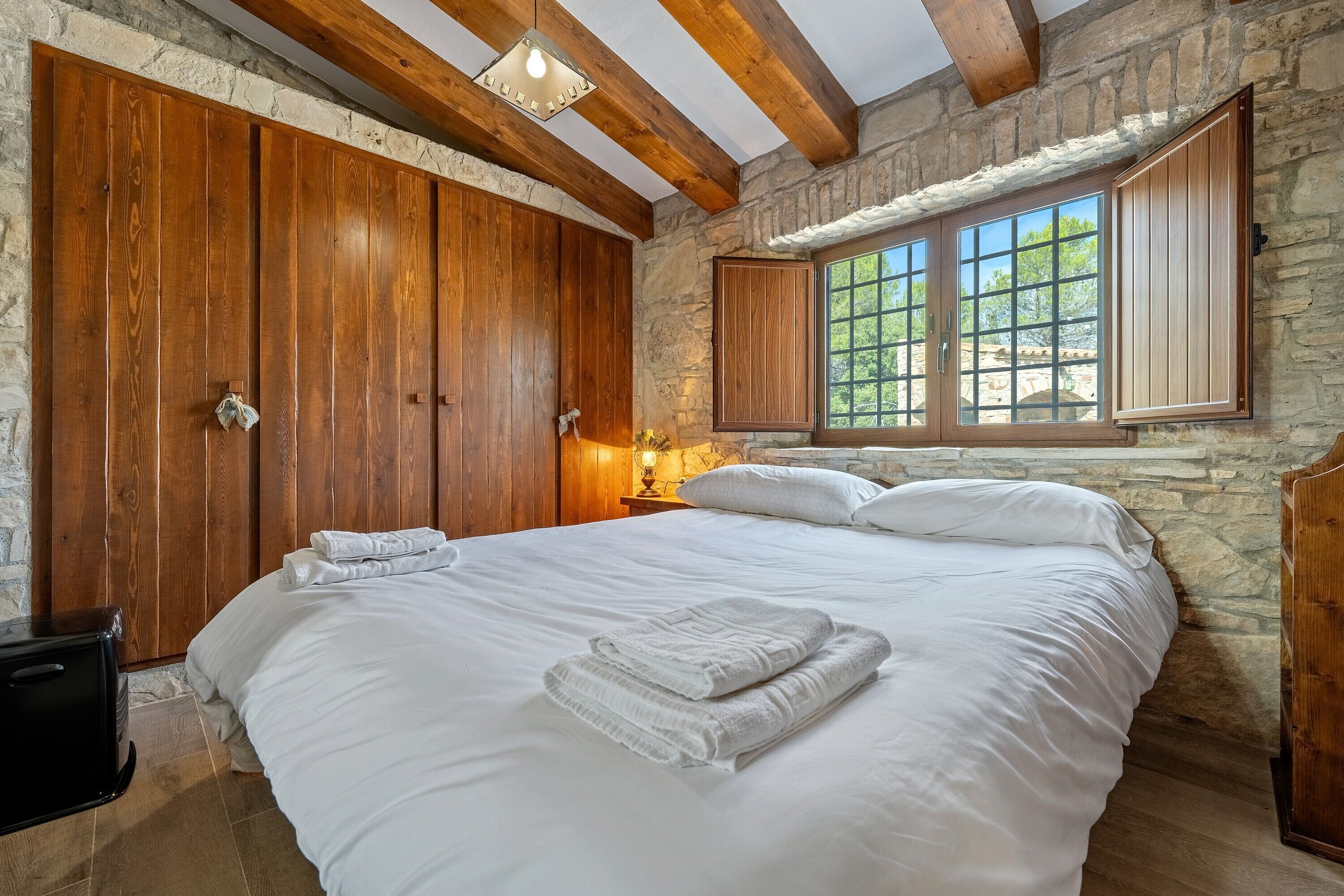 A bedroom with a large bed, wooden wardrobe, stone walls, and a window with shutters.