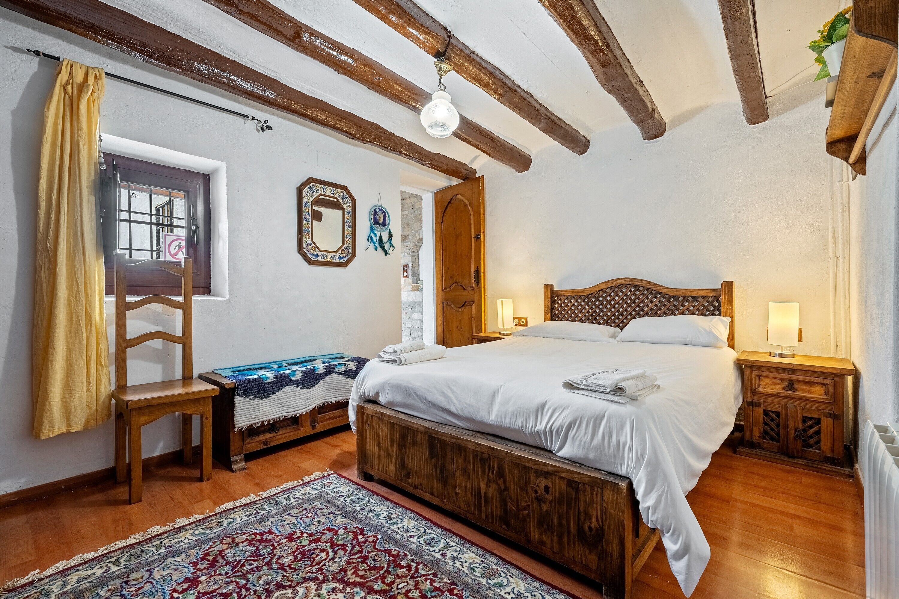 A bedroom with a wooden bed, a nightstand, a chair, a window with curtains, and a patterned rug.