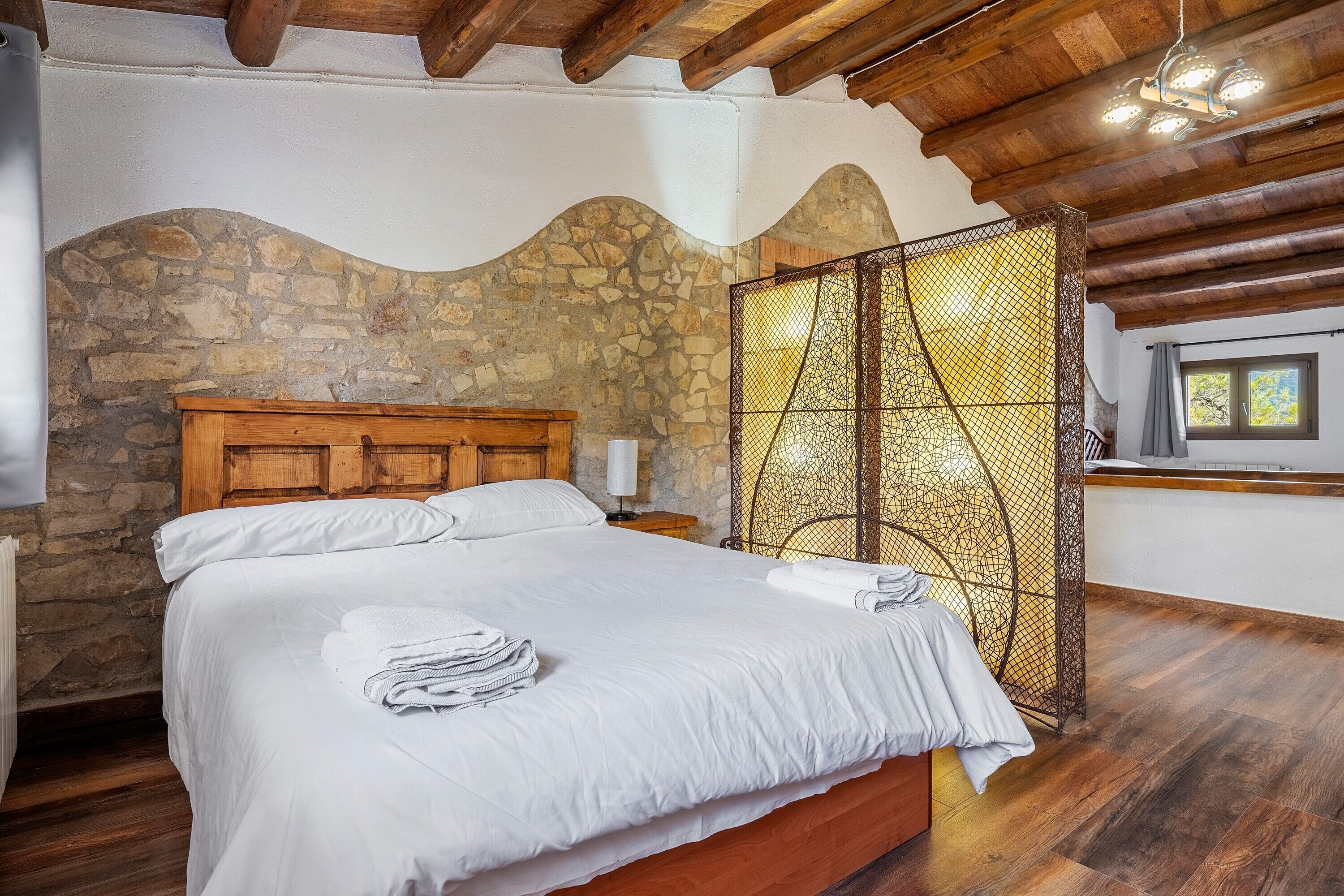 Une chambre avec un mur en pierre, des poutres en bois au plafond, un lit avec du linge de lit blanc, une table de chevet et une fenêtre avec des rideaux.