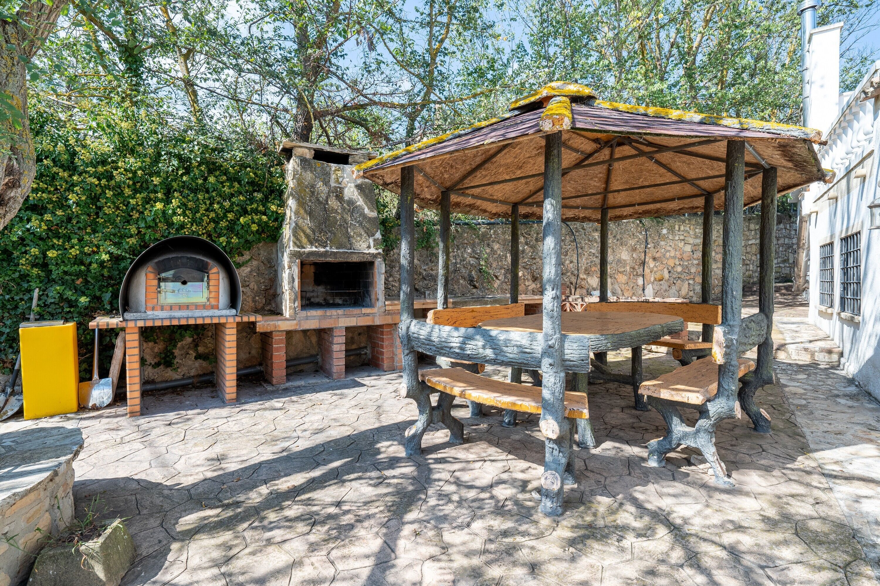An outdoor patio with a wooden table and benches, a brick oven, and a covered seating area.