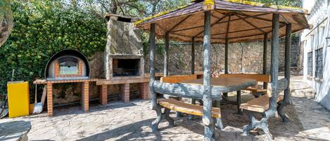 An outdoor patio with a wooden table and benches, a brick oven, and a covered seating area.