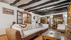 A traditional interior with exposed wooden beams, a wicker sofa, a wooden table, and a rustic kitchen area. - Chalet "Can Tronc" with Mountain View, Shared Pool & Wi-Fi (Querol)