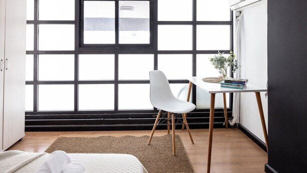 A room with a large window, a white chair, a small table with books, and a bed with a white blanket.