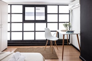 Une pièce avec une grande fenêtre, une chaise blanche, une petite table avec des livres et un lit recouvert d’une couverture blanche.