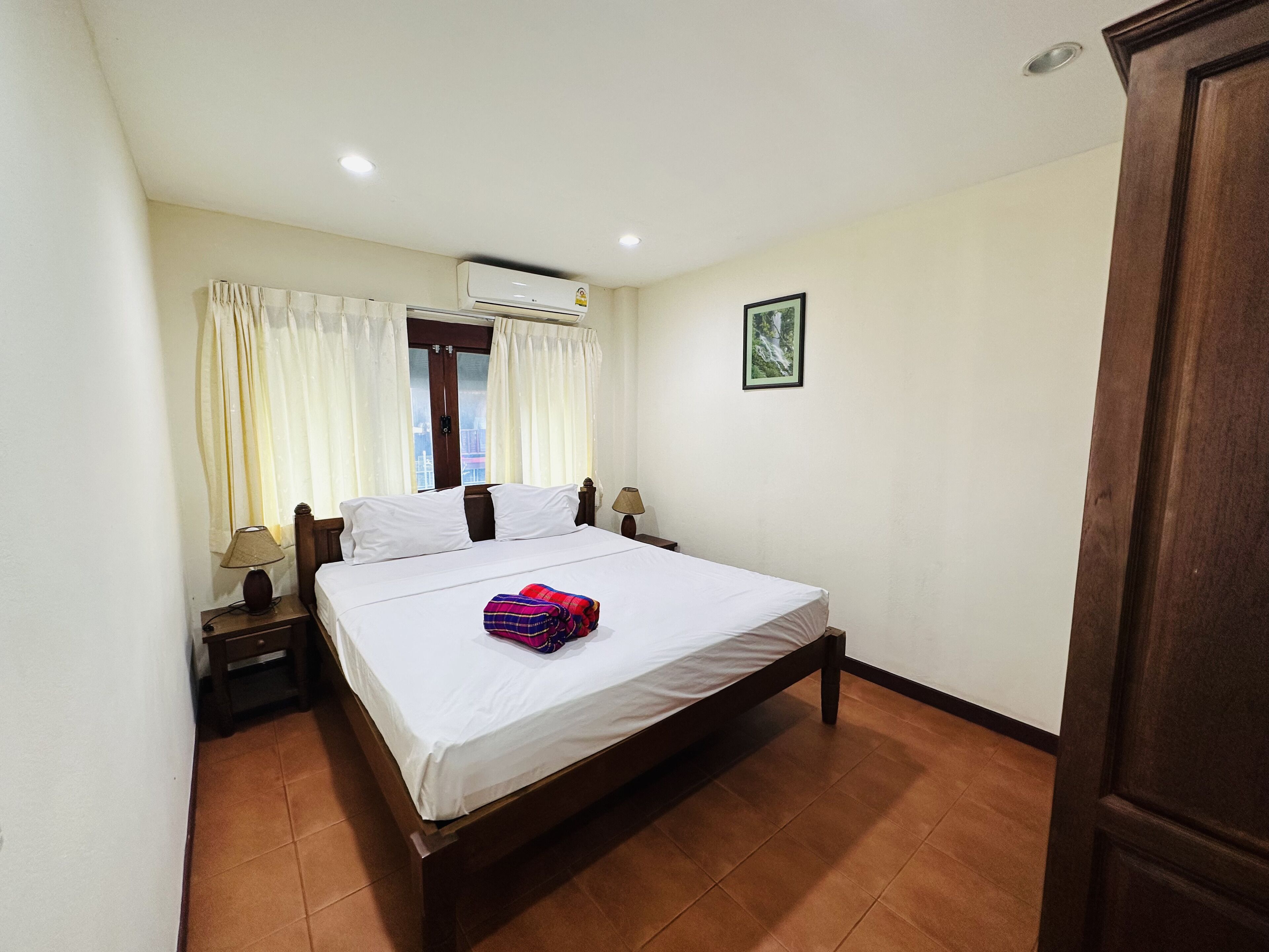 A hotel room with a bed, curtains, an air conditioning unit, a nightstand, a lamp, a framed picture on the wall, and a door.