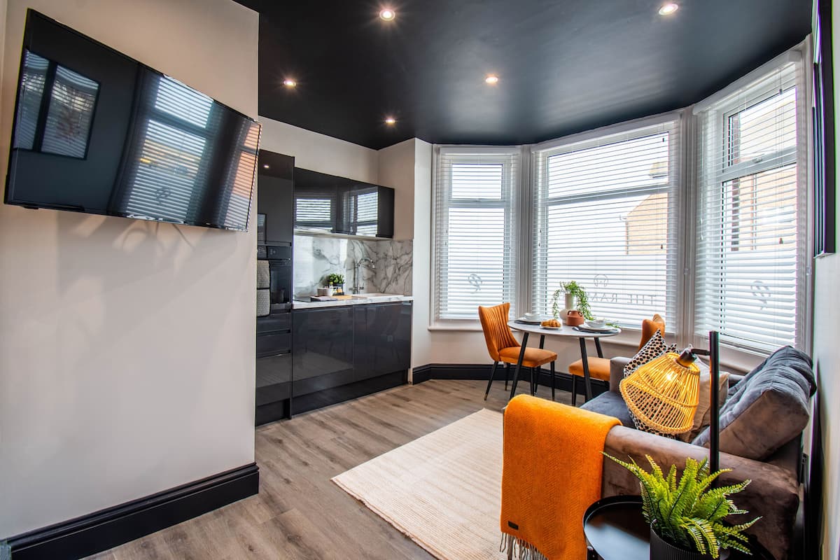 A modern kitchen with dark cabinets and a marble countertop, a dining area with a table and chairs, and a cozy seating area with a sofa and a potted plant.
