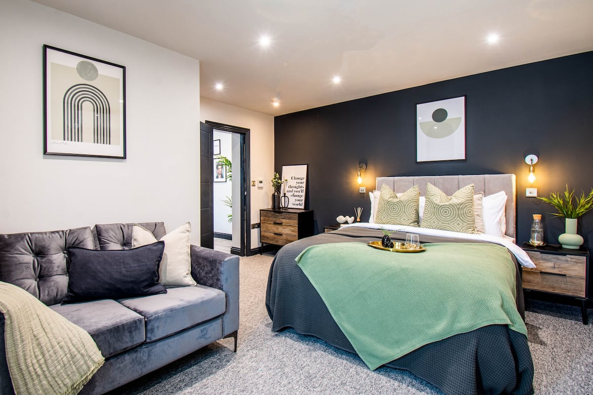 A modern bedroom with a grey sofa, a large bed with patterned pillows, and a green throw blanket.