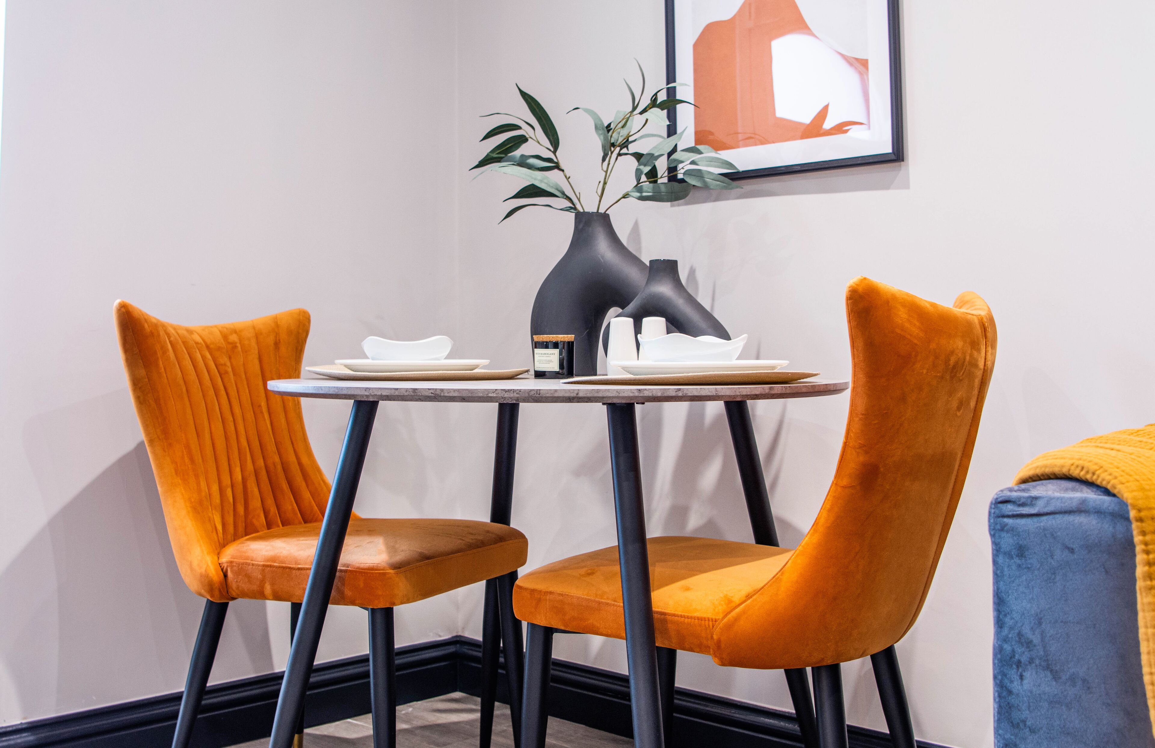 Un comedor con una mesa redonda, dos sillas anaranjadas, un jarrón con plantas verdes y una obra de arte enmarcada en la pared.