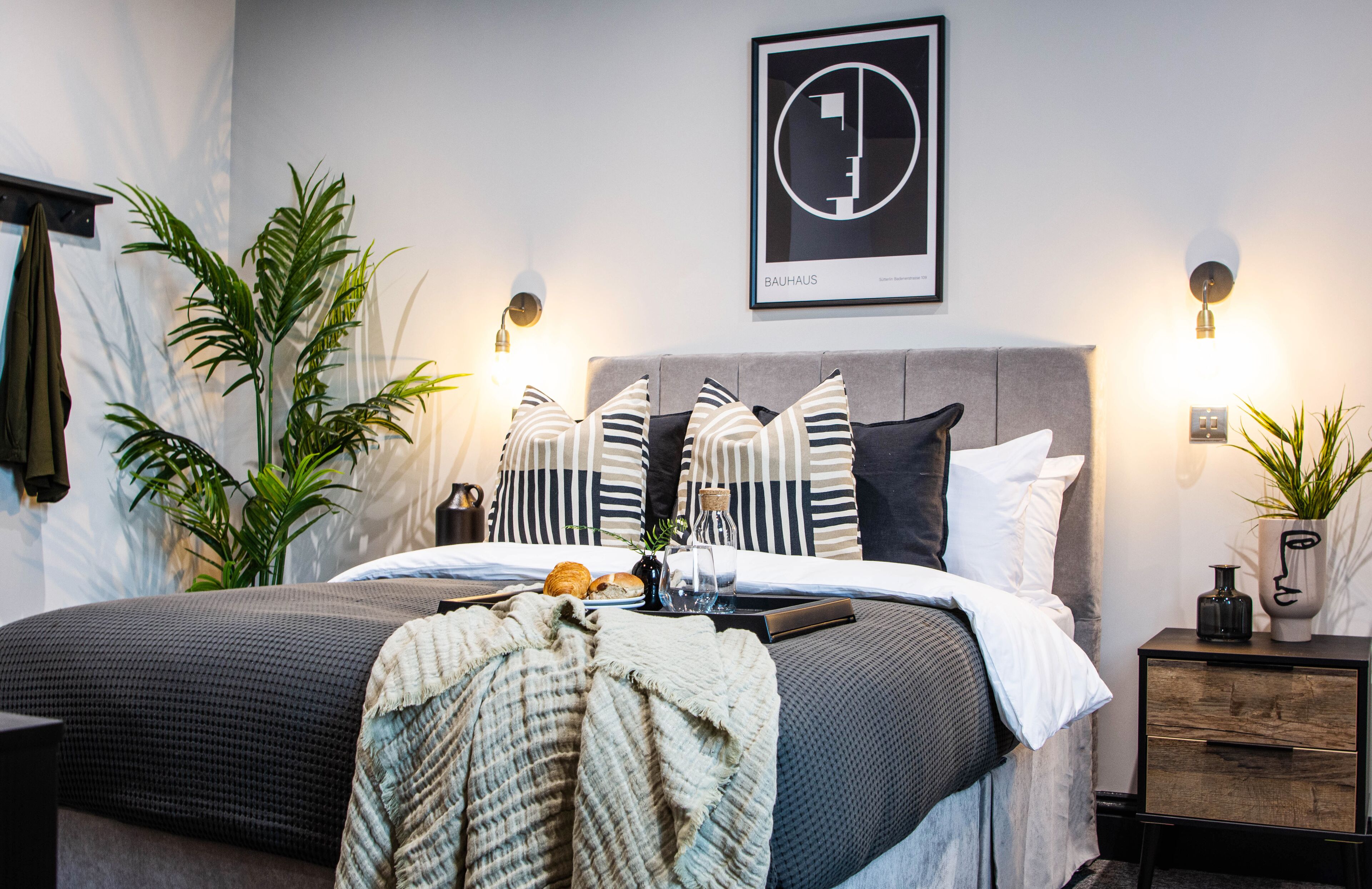 Ein modernes Schlafzimmer mit grauem Bett, gestreiften Kissen und einem Bauhaus-Kunstwerk in einem Rahmen an der Wand.