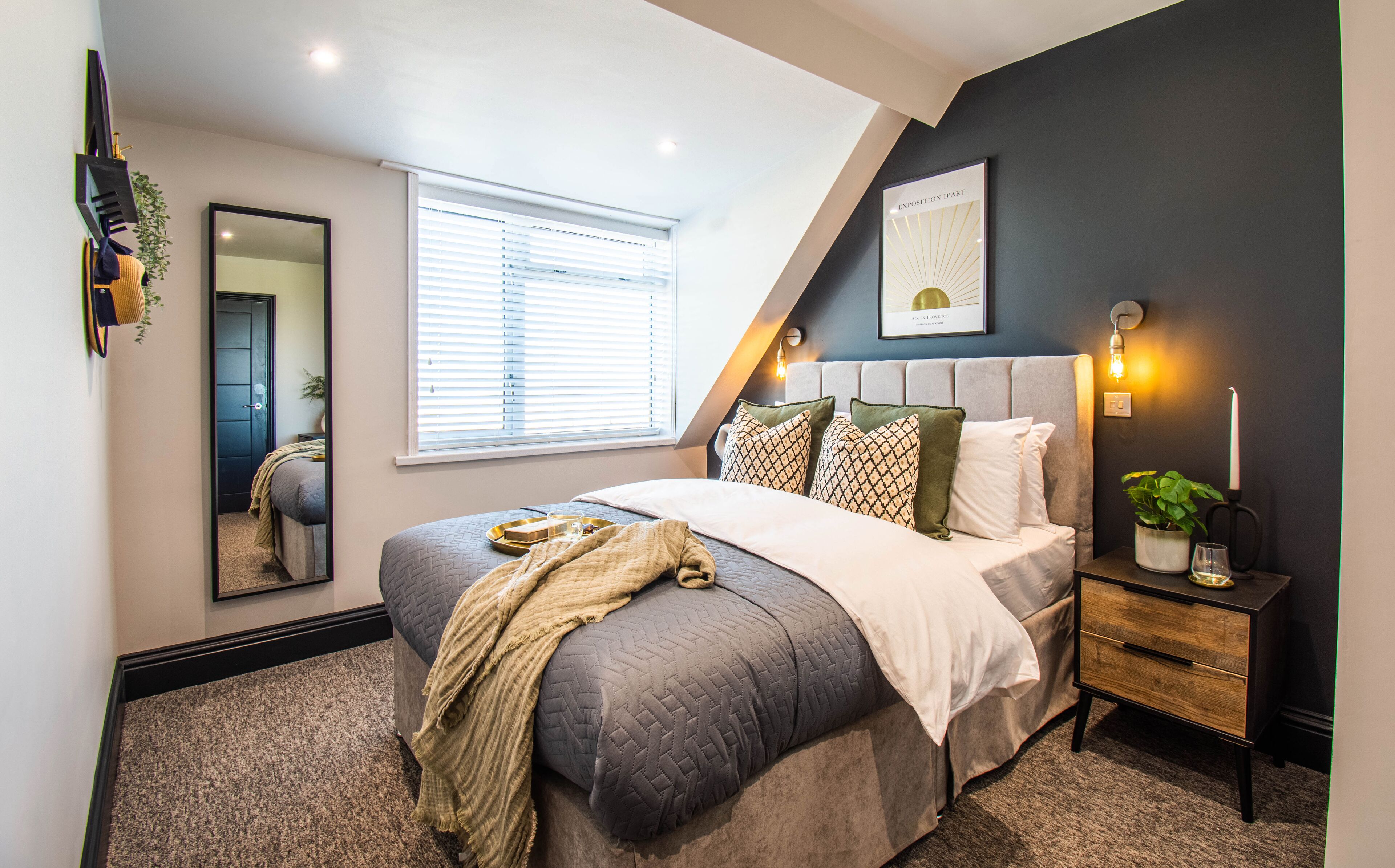 A modern bedroom with a large bed, a bedside table, a nightstand, a mirror, a window with blinds, and a framed picture on the wall.