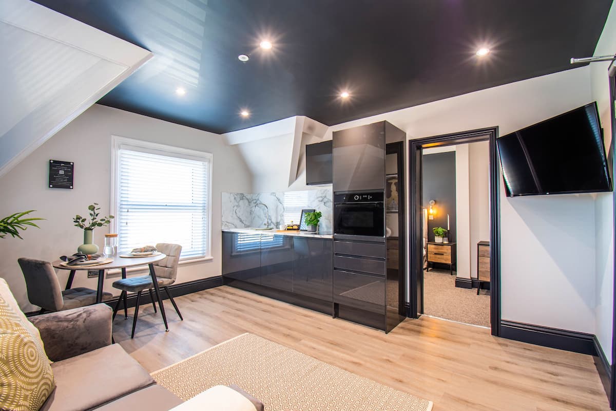 A modern kitchen with a black refrigerator, a microwave, and a stove. There is a dining area with a table and chairs, and a large window with blinds.