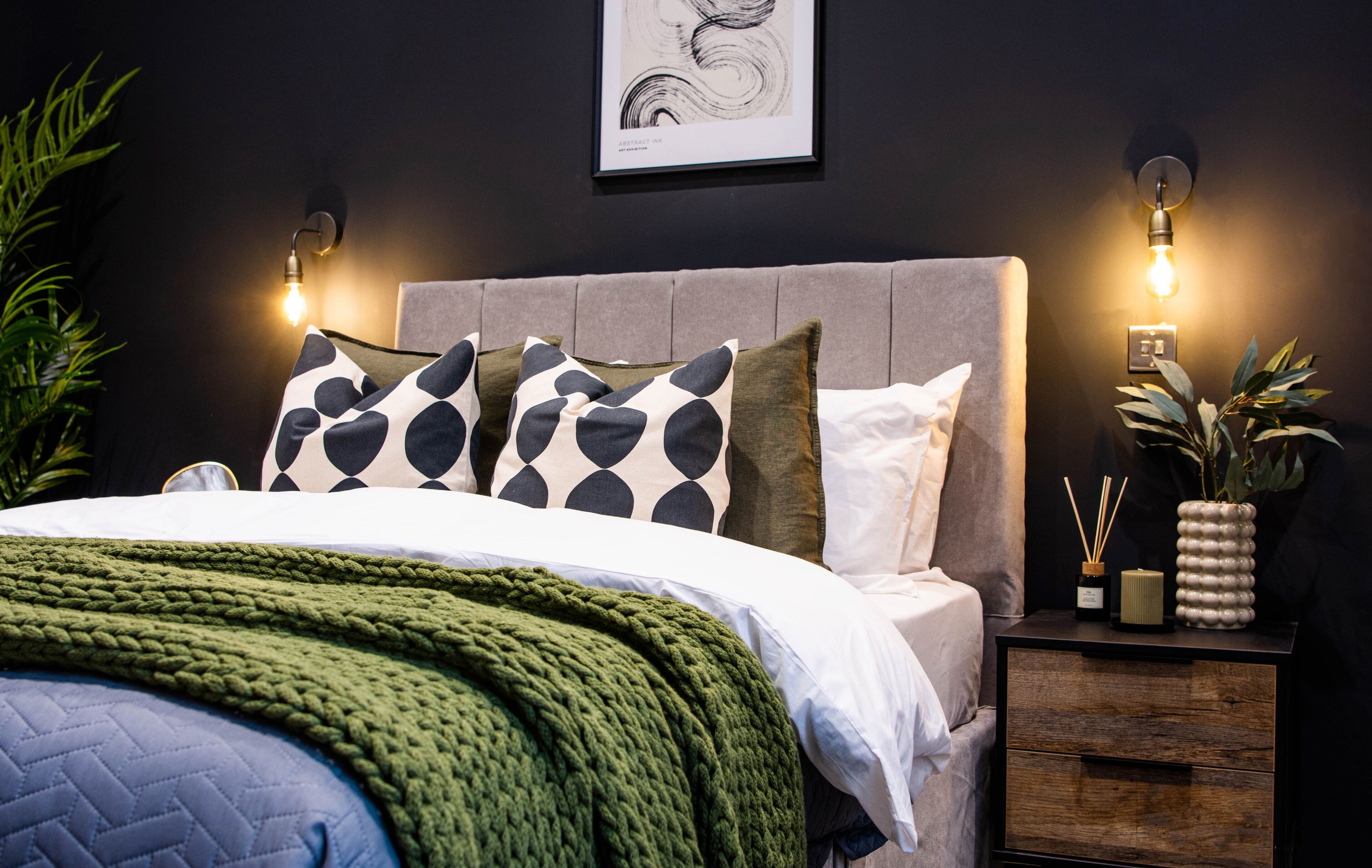 Un lit bien fait, recouvert d’une couverture verte, avec une tête de lit grise et des coussins à motifs. Des lampes murales et une œuvre d’art encadrée sont disposées au-dessus du lit.