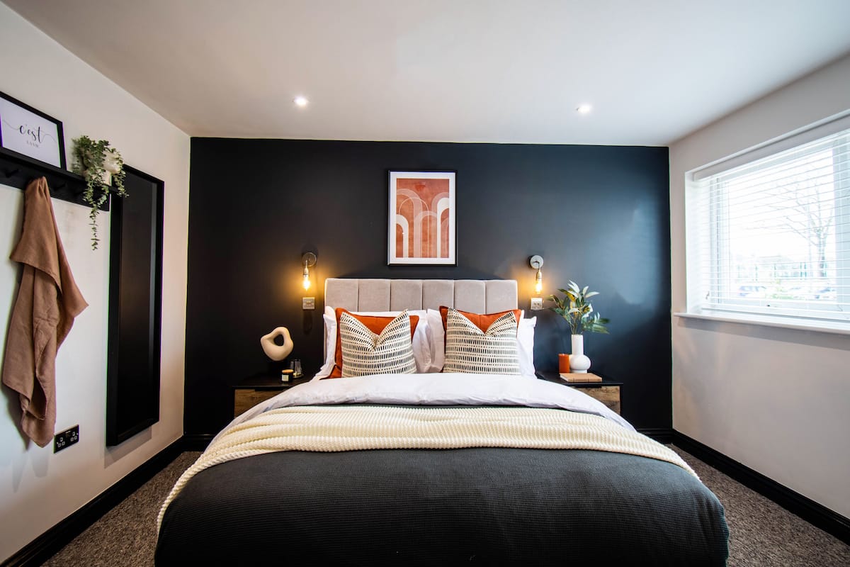 A modern bedroom with a large bed, a dark accent wall, a framed artwork above the bed, and a window with blinds.