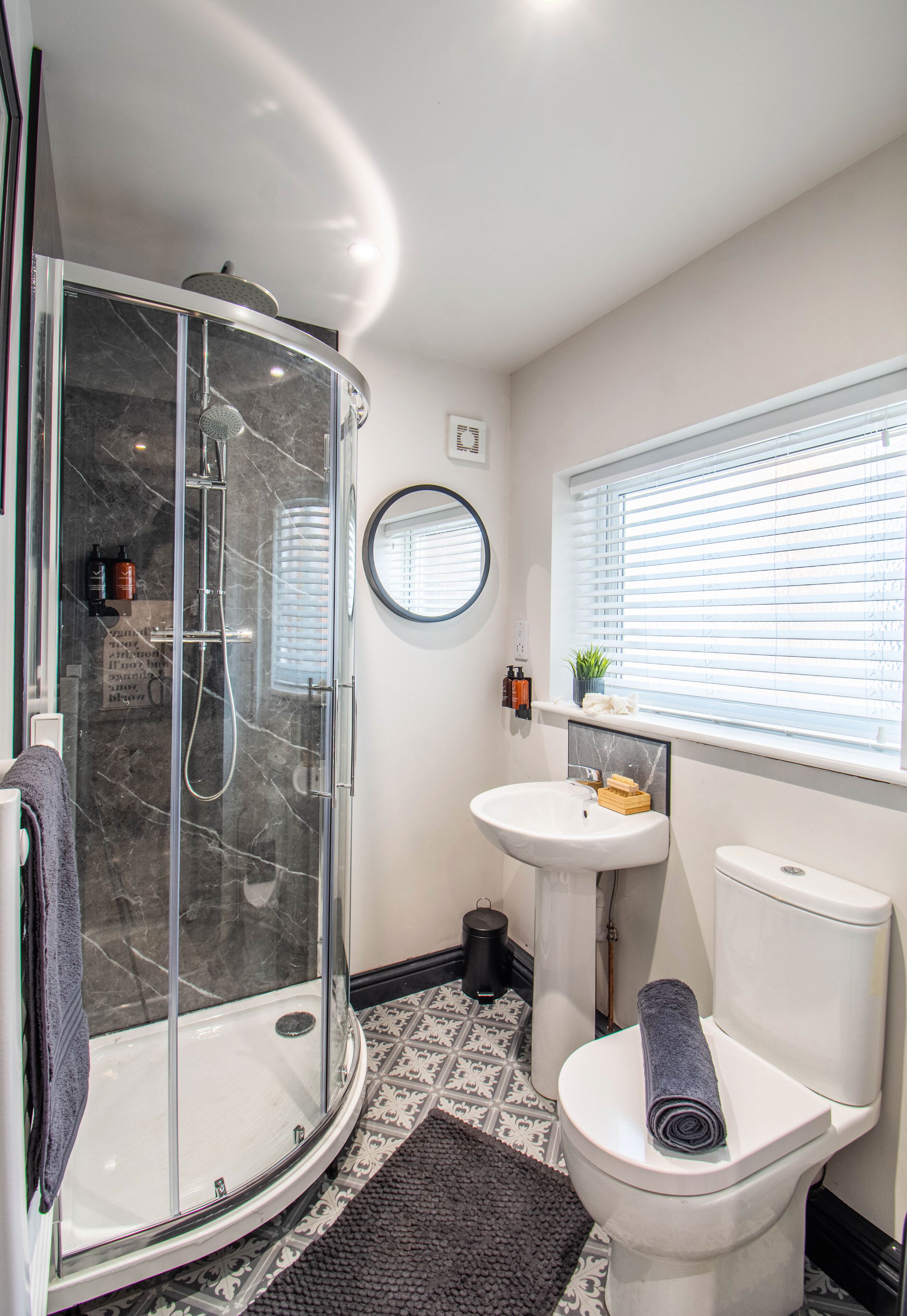 A bathroom with a glass-enclosed shower, a pedestal sink, a toilet, and a window with blinds.