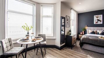 A modern bedroom with a bed, bedside tables, a chair, a small table with a vase and a bowl of fruit, and large windows with blinds.