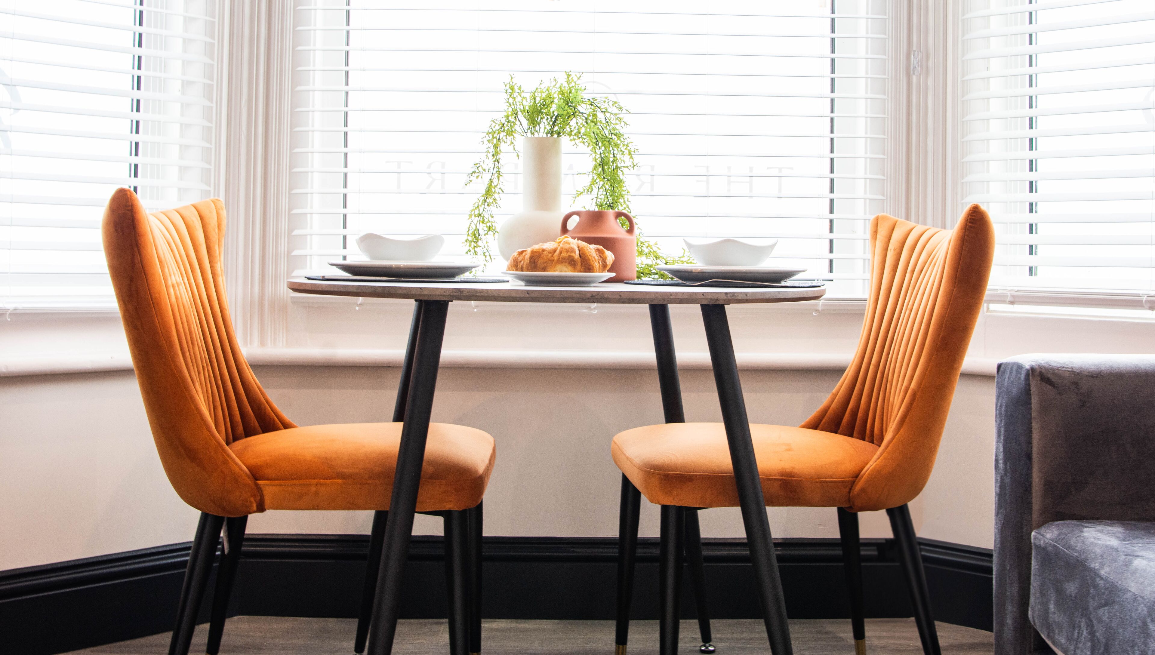 A dining area with two orange chairs, a small table, and a window with blinds.