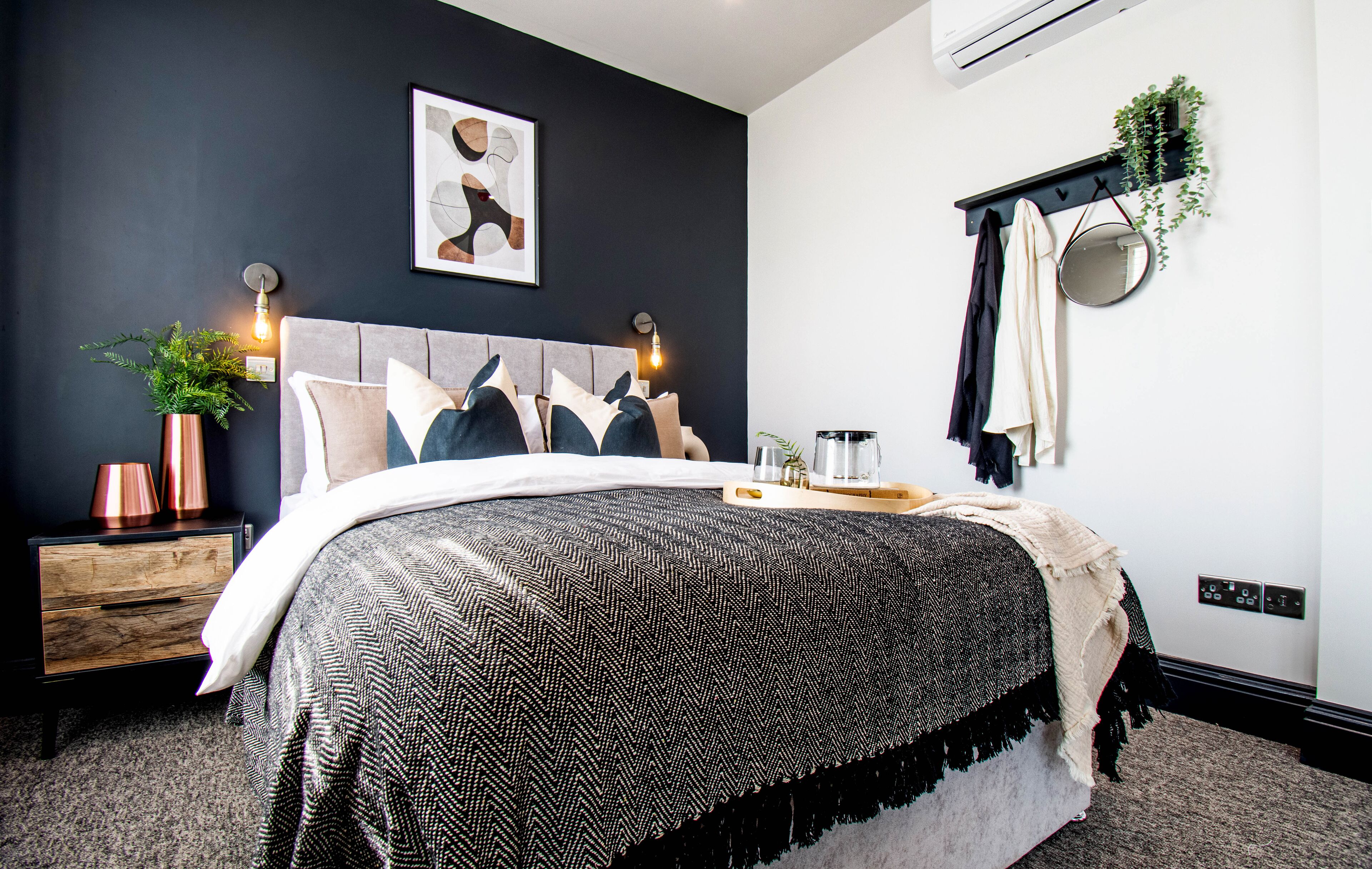 A modern bedroom with a dark wall, a grey headboard, and a bed with a patterned bedspread. There is a bedside table with a lamp and a plant.