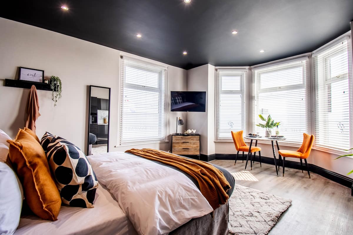 A modern bedroom with a bed, a TV, a desk with two chairs, and large windows with blinds.