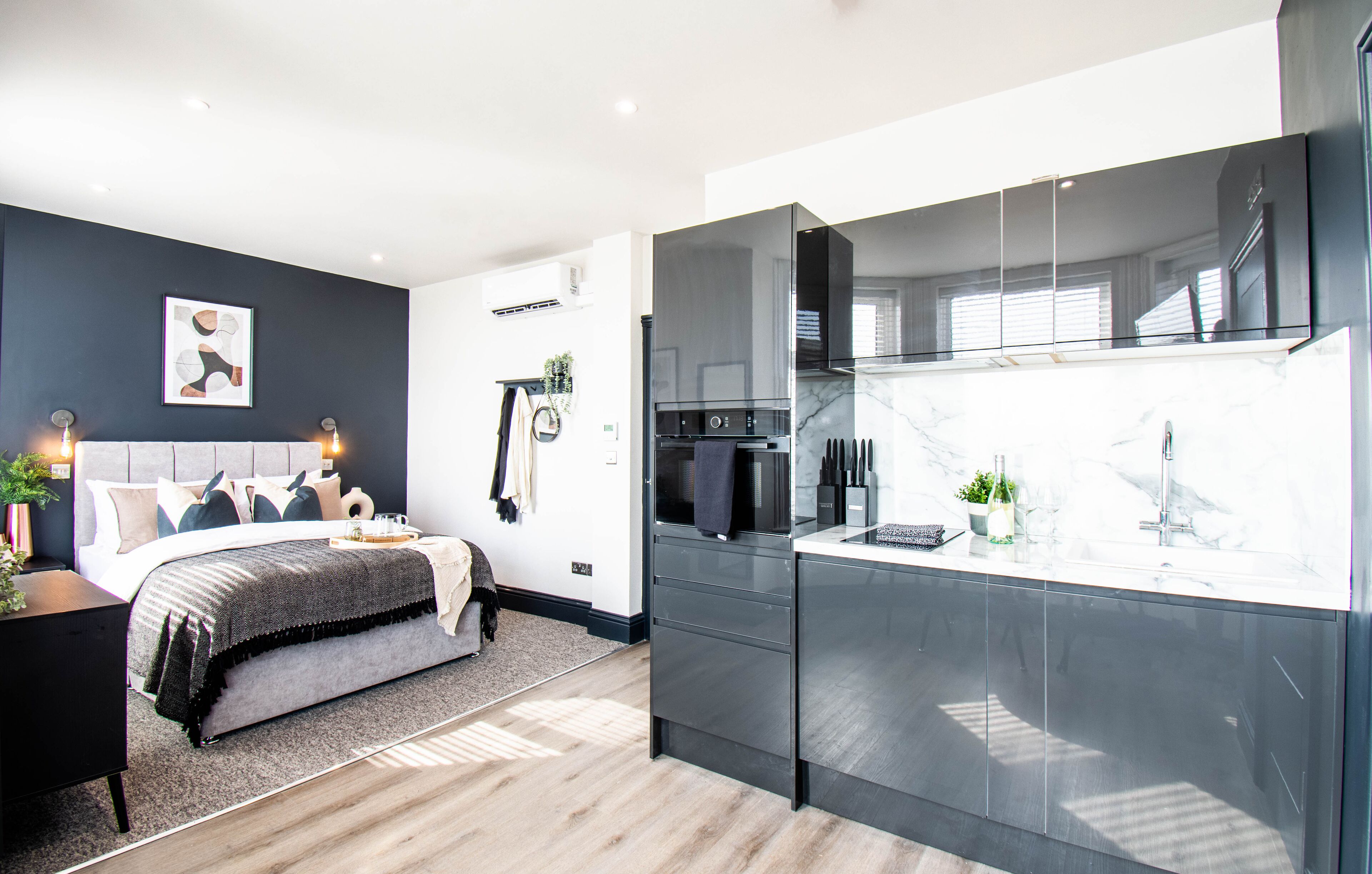 Ein modernes Schlafzimmer mit einem großen Bett, Nachttischen und einem kleinen Tisch daneben. Angrenzend an das Schlafzimmer befindet sich eine zeitgemäße Küche mit einer großen Kochinsel, einem eingebauten Backofen und einer Glasrückwand.