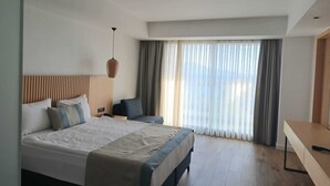 A modern hotel room with a large bed, a desk, and a view of the outdoors through sheer curtains. - Ömer Prime Holiday Resort (Kusadasi)