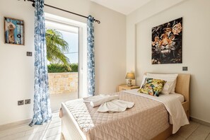 A bedroom with a bed, a nightstand, a lamp, a painting, and a view of the outdoors. - Enjoy your vacation at a luxurious stone villa with garden & swimming pool! (Agioi Theodoroi)