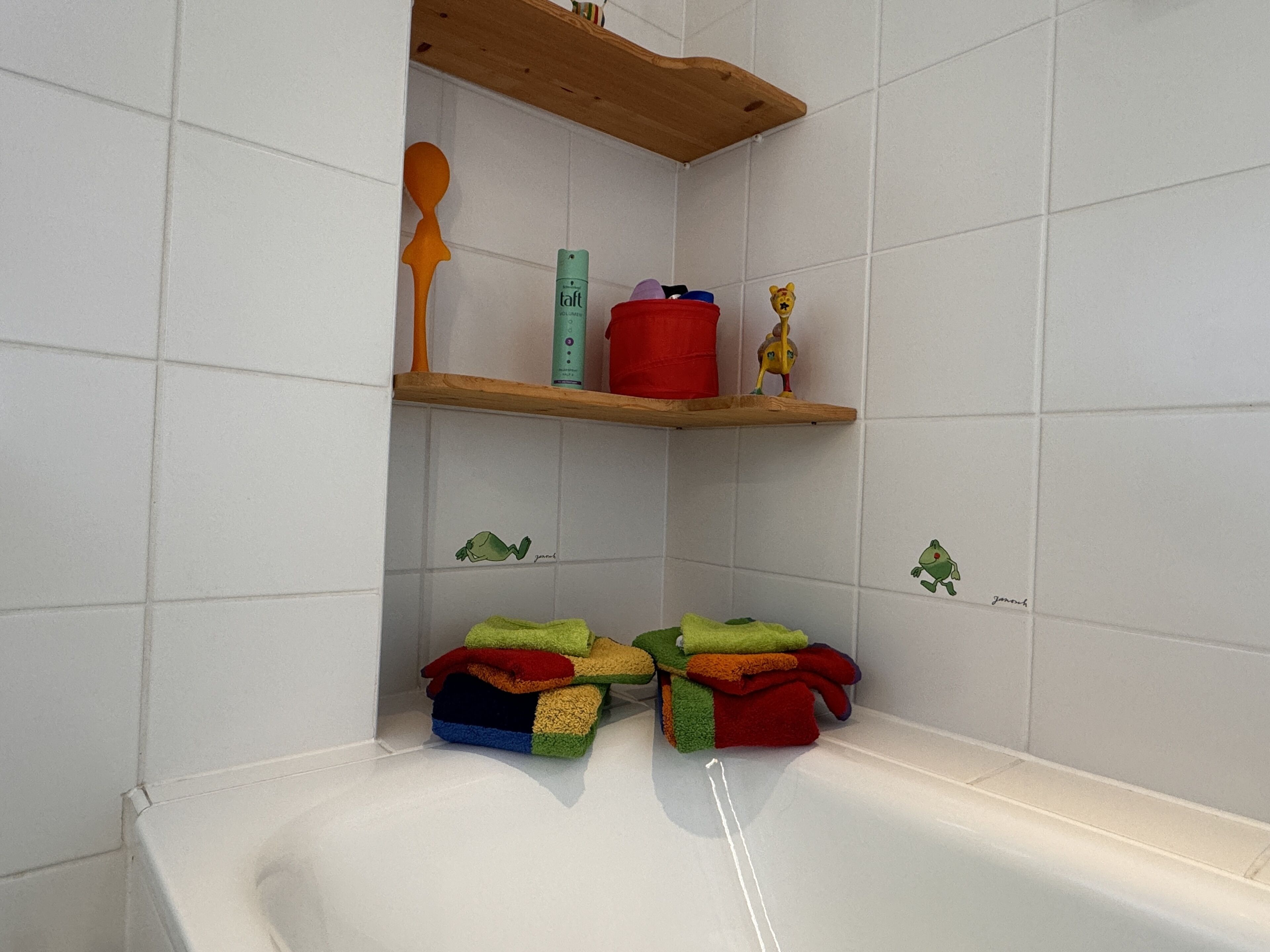 A bathroom with white tiles, a wooden shelf with toiletries, and a stack of colorful towels.