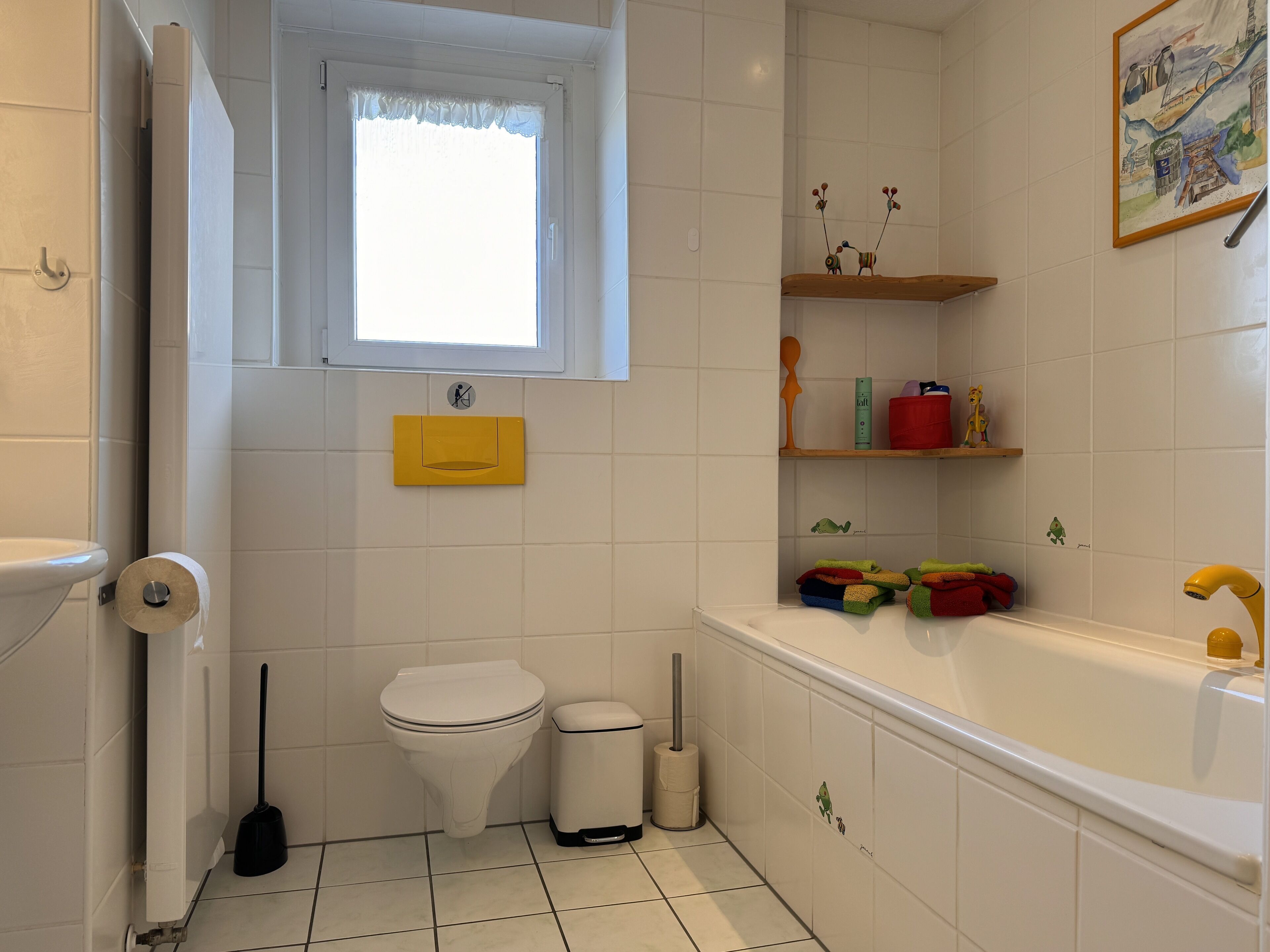 Un baño con inodoro, bidé y bañera. Hay una ventana con cortina, un estante con artículos de aseo y un cuadro en la pared.