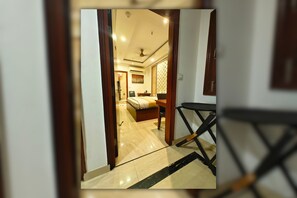 A hotel room with a bed, a desk, and a chair. There is a door leading to another room. - Hotel MGM Residency (New Delhi)