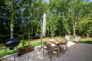 A picnic setup with a grill, wooden table, and chairs in a forested area. - Cottage "Cocooning" (Saint-Chamassy)