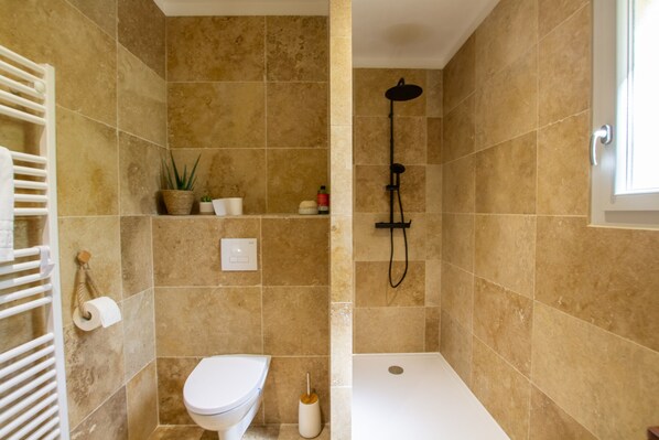 Un baño con baldosas color beige, un inodoro blanco, una ducha con cabezal de ducha manual, un perchero y un estante pequeño con artículos de aseo.