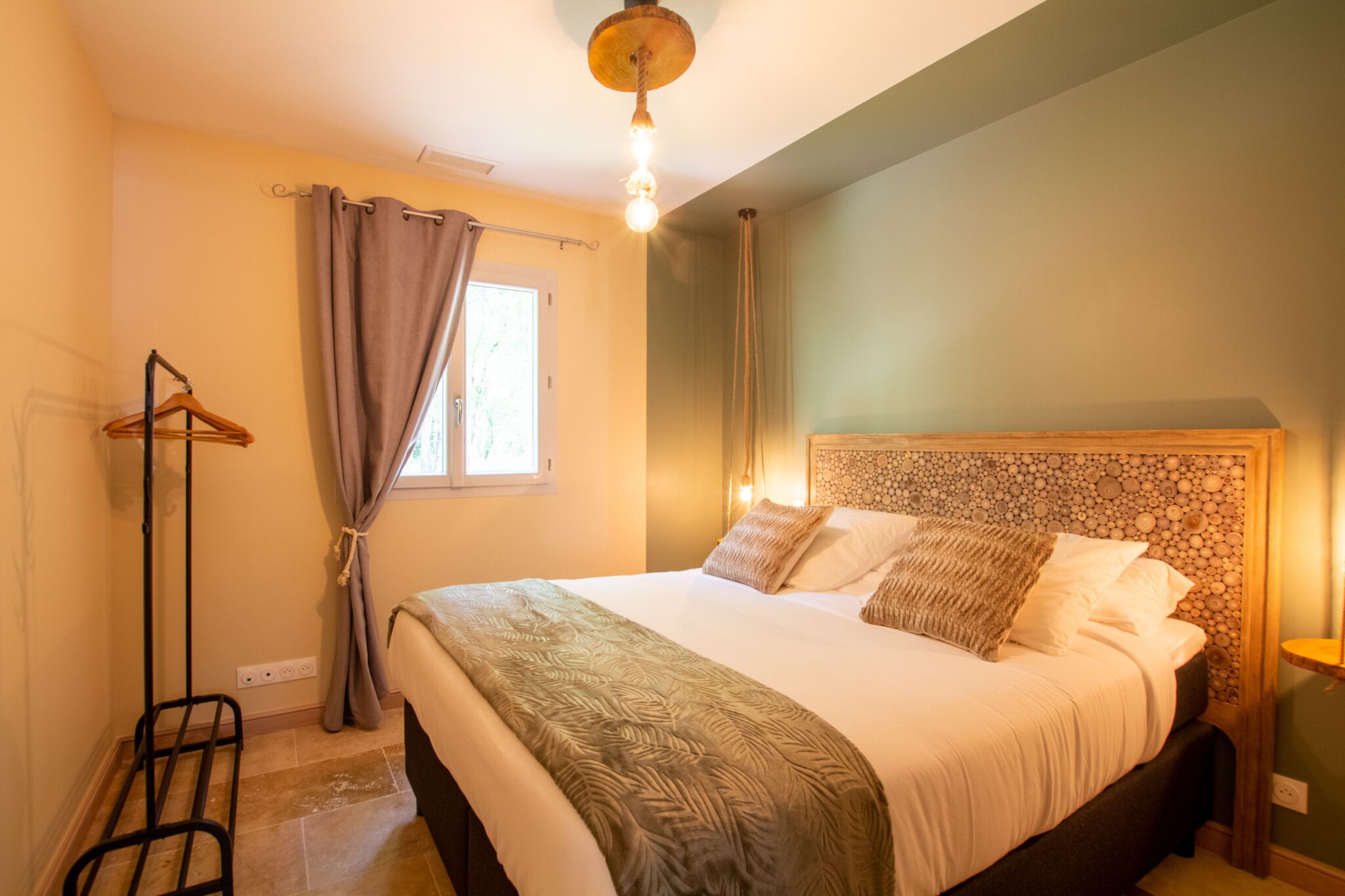 Une chambre à coucher avec un grand lit, une tête de lit en bois, un luminaire suspendu et une fenêtre avec des rideaux.