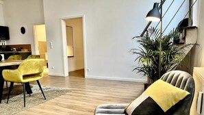 A modern living room with a wooden floor, a grey armchair with a yellow pillow, a yellow sofa, and a dining area with a table and chairs.