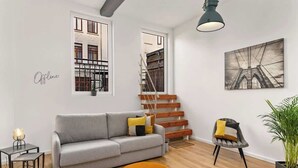 A modern living room with a grey sofa, a wooden staircase, a black and white artwork, and a plant. - MEL&BENS Loft 2 | Terrace | Design | Weserstadion (Bremen)
