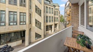A balcony with a wooden table and a potted plant, overlooking a street with buildings. - MEL&BENS Domshof | City center | Balcony | (Bremen)